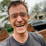 Smiling young man with muddy face and glasses standing outdoors in a casual t-shirt.